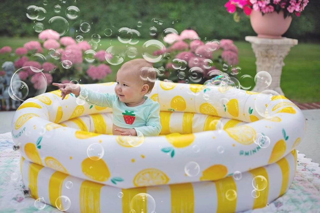 Un bébé assis dans une piscine gonflable jaune rayée, entouré de bulles de savon, dans un jardin avec fleurs roses