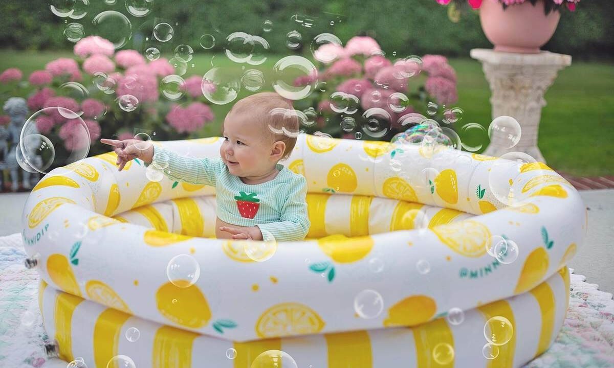 Un bébé assis dans une piscine gonflable jaune rayée, entouré de bulles de savon, dans un jardin avec fleurs roses