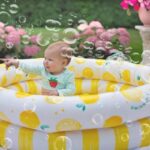 Un bébé assis dans une piscine gonflable jaune rayée, entouré de bulles de savon, dans un jardin avec fleurs roses