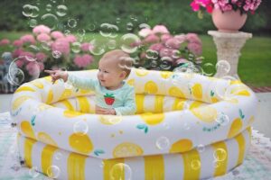 Un bébé assis dans une piscine gonflable jaune rayée, entouré de bulles de savon, dans un jardin avec fleurs roses