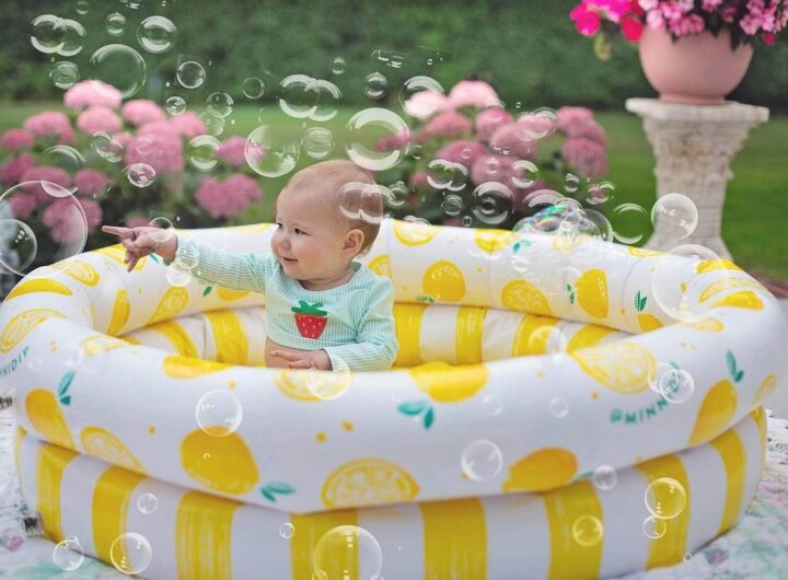 Un bébé assis dans une piscine gonflable jaune rayée, entouré de bulles de savon, dans un jardin avec fleurs roses